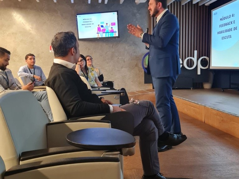 Palestrante de terno conduz o módulo de feedback e escuta em auditório corporativo, diante de participantes sentados em poltronas