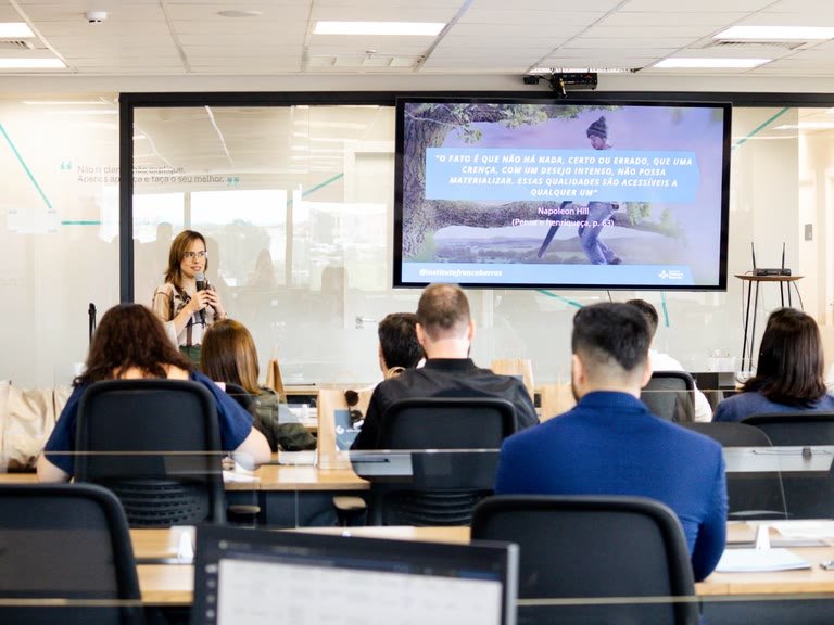 Treinamento em sala de reunião corporativa: a palestrante fala ao microfone enquanto o telão exibe um slide do Instituto França Barros